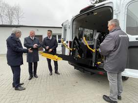 Pokaż zdjęcie: Z tyłu samochodu stoi trzech mężczyzn, którzy rozmawiają ze sobą. Jest opuszczana winda umożliwiająca wjazd do wnętrza auta wózkom inwalidzkim. 