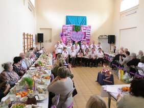 Pokaż zdjęcie: Na środku sali stoi grupa występujących artystów. Po bokach są ustawione stoły, przy których siedzą ludzie. Cała sala jest udekorowana świątecznymi ozdobami. 