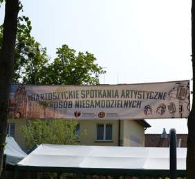 Pokaż zdjęcie: Baner z nazwą wydarzenia i panoramą Bartoszyc zawieszony na drzewach. Na drugim planie budynki mieszkalne.