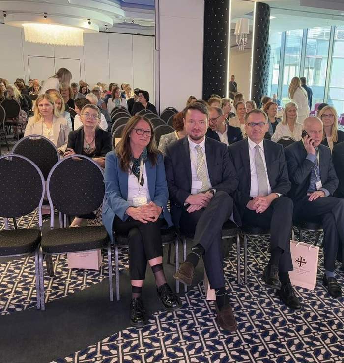 Pokaż zdjęcie: Grupa uczestników siedzi w pierwszych rzędach sali konferencyjnej. Na pierwszym planie widać pięć osób ubranych w formalne stroje, siedzących na czarnych krzesłach z torbami konferencyjnymi u stóp. W tle widać pozostałych uczestników i nowoczesne wnętrze z lustrzaną ścianą i żyrandolem. Panuje atmosfera skupienia i oczekiwania na rozpoczęcie wydarzenia.