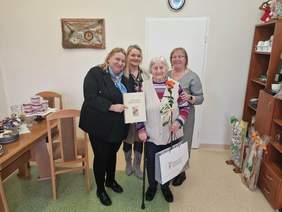 Pokaż zdjęcie: Na zdjęciu znajdują się cztery uśmiechnięte i ubrane odświętnie kobiety, a wśród nich jedna w wieku starszm, która trzyma nagrodę PFRON