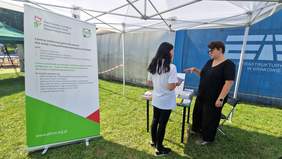 Pokaż zdjęcie: Plenerowe stoisko informacyjne PFRON. obok stolika z ulotkami stoi rollup z logo PFRON. Ekspertka PFRON udziela informacji kobiecie, która podeszła do stolika.