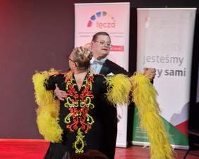 Pokaż zdjęcie: Dwie osoby z zespołem Downa tańczą na scenie. Ubrani są w profesjonalne stroje do tańca towarzyskiego.