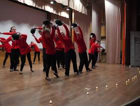 Pokaż zdjęcie: Zdjęcie przedstawia występ grupy osób na scenie. Osoby są ubrane w czerwone bluzy i czarne spodnie, a w rękach trzymają czarne kapelusze uniesione nad głowami jak w układzie tanecznym. Scena ma drewnianą podłogę, a z przodu na podłodze stoi rząd małych świeczek. W tle widać zasłony i oświetlenie sceniczne.