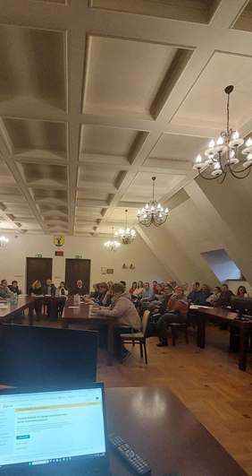 Pokaż zdjęcie: Na pierwszym planie laptop, na drugim kilkadziesiąt osób siedzących przy długich stołach.