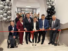 Pokaż zdjęcie: Grupa sześciu elegancko ubranych osób przecina czerwoną wstęgę podczas uroczystego otwarcia; po bokach duże czarno-srebrne dekoracje z balonów, za nimi stoi tłum gości obserwujących wydarzenie.