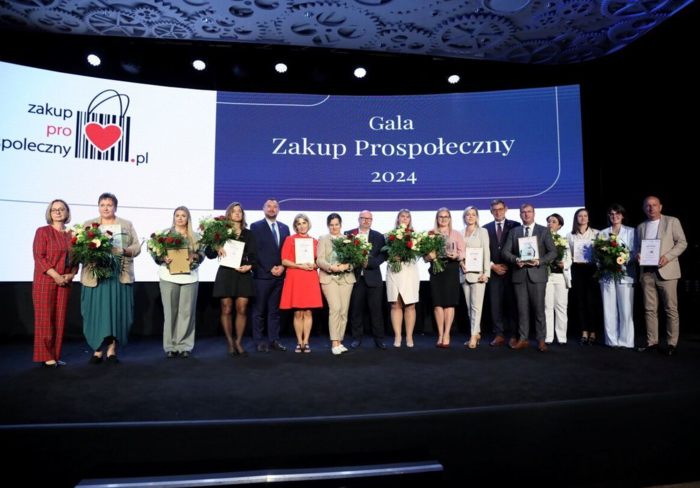 Pokaż zdjęcie: Na scenie, na tle napisu Gala zakup prospołeczny stoi grupa siedemnastu osob, pozując do pamiątkowego zdjęcia. Większość osób trzyma kwiaty i dyplomy.