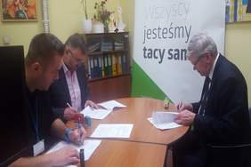 Pokaż zdjęcie: pomieszczenie biurowe w siedzibie Oddziału Łódzkiego PFRON. Przy stole siedzą  trzej mężczyźni i podpisują dokumenty dotyczące dofinansowania do oprocentowania kredytów bankowych 