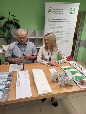 Pokaż zdjęcie: Przy biurku na którym leżą ulotki i katalogi siedzi mężczyzna i  kobieta. W tle baner z logo CIDON.