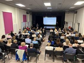 Pokaż zdjęcie: Duża grupa osób siedzi w sali konferencyjnej i słucha wystąpienia kobiety, która stoi po lewej stronie od sceny. W sali ustawione zostały również banery organizatorów wydarzenia. Na dużym ekranie wyświetlana jest prezentacja, a na scenie znajduje się tłumacz języka migowego.