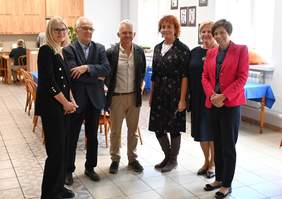 Pokaż zdjęcie: Grupa sześciu osób stoi w sali, w której znajdują się stoły z krzesłami. W tle widoczne są szafki podwieszone na jednej ze ścian oraz portrety zawieszone na drugiej ścianie.