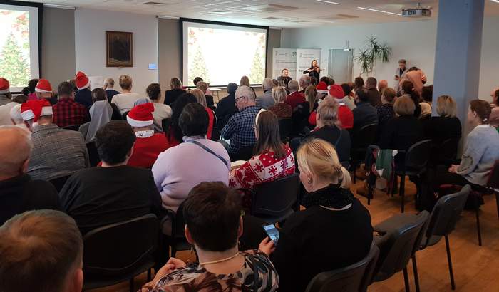 Pokaż zdjęcie: Duża grupa osób siedzi w sali konferencyjnej. Część z tych osób nosi czapkę Świętego Mikołaja. W sali są dwa ekrany, na których widać przyozdobione choinki. Po prawej stronie znajdują się dwa banery PFRON oraz występuje duet instrumentalistów.