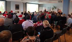 Pokaż zdjęcie: Duża grupa osób siedzi w sali konferencyjnej. Część z tych osób nosi czapkę Świętego Mikołaja. W sali są dwa ekrany, na których widać przyozdobione choinki. Po prawej stronie znajdują się dwa banery PFRON oraz występuje duet instrumentalistów.