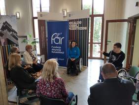 Pokaż zdjęcie: 5 osób siedzi w kole na korytarzu 2 osoby kobieta i mężczyzna siedzą na wózkach inwalidzkich obok stoi baner z białym napisem na granatowym tle fundacja aktywnej rehabilitacji