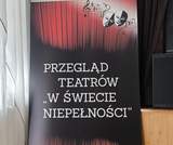Roll-up z napisem „Przegląd Teatrów «W Świecie Niepełnosprawności»”, z grafiką czerwonych kurtyn, masek teatralnych i nut. Baner ustawiony w sali obok zasłony i głośników