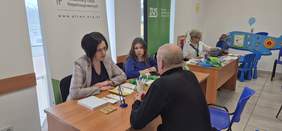 Pokaż zdjęcie: Przy biurku na którym leżą ulotki, siedzą dwie kobiety udzielające informacji mężczyźnie.