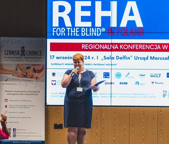 Pokaż zdjęcie: Prowadząca na scenie w niebieskiej sukience, w tle ekran z infografiką na temat konferencji