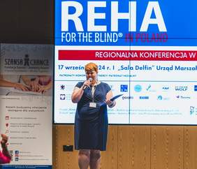 Pokaż zdjęcie: Prowadząca na scenie w niebieskiej sukience, w tle ekran z infografiką na temat konferencji