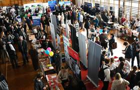 Widok z góry na Targi Pracy w sali edukacyjnej – liczne stoiska instytucji i firm, roll-upy informacyjne, osoby odwiedzające rozmawiają z wystawcami i zapoznają się z ofertami pracy oraz materiałami promocyjnymi.