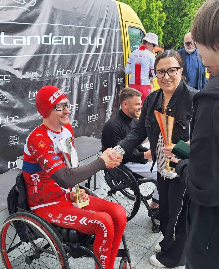 Pokaż zdjęcie: Kobieta w czarnych okularach wręcza puchar zawodnikowi siedzącemu na wózku inwalidzkim. W lewej ręce kobieta trzyma puchar.