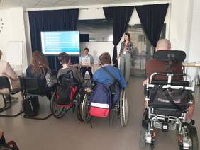 Na zdjęciu widzimy grupę uczestników warsztatów siedzących na krzesłach lub wózkach inwalidzkich, skierowanych w stronę ekranu, na którym wyświetlane są slajdy z prezentacji. W tle prowadzący przedstawiają treści dotyczące dostępności.