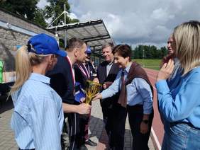 Pokaż zdjęcie: Grupa pięciu osób, kobieta w niebieskiej bluzce gratuluje mężczyźnie w garniturze, ściskając mu rękę.