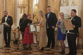 Pokaż zdjęcie: W sali wielkiej Zamku Królewskiego w Warszawie laureatka odbiera nagrodę, w tle stoją inni laureaci. 