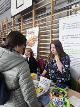 Pokaż zdjęcie: Stoisko PFRON z materiałami informacyjnymi i publikacjami. Osoba stojąca rozmawia z osobą siedzącą przy stole. W tle widoczny roll-up z informacjami o Centrach Informacyjno‑Doradczych dla Osób z Niepełnosprawnością.