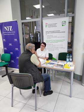 Pokaż zdjęcie: Kobieta reprezentująca PFRON rozmawia przy stole z mężczyzną podczas wydarzenia informacyjnego. Na stole znajdują się ulotki, broszury i woda. W tle widoczne są banery PFRON i NFZ.