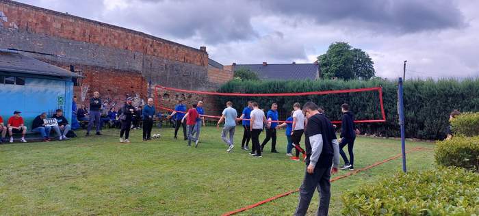 Pokaż zdjęcie: Dwie drużyny znajdują się na boisku po obu stronach siatki i idąc, witają się rękoma. Przy budynku z cegły znajdują się kibice oraz sędzia.