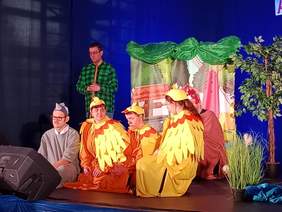 Pokaż zdjęcie: Na scenie siedzą aktorzy. Są przebrani za zwierzęta. Jest także sztuczne drzewo i trawa. 