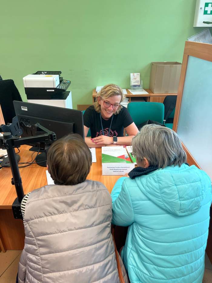 Pokaż zdjęcie: 2 kobiety siedzą przed biurkiem, a pracownik PFRON udziela informacji