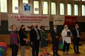 Pokaż zdjęcie: Fragment sali gimnastycznej w rzędzie stoi 7 osób 3 kobiety i 4 mężczyzn w tle baner z napisem śląski dzień treningowy programu aktywności motorycznej olimpiad specjalnych