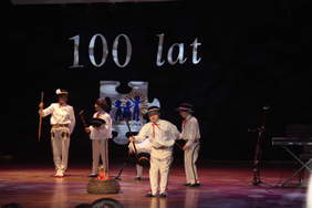 Pokaż zdjęcie: Na scenie czterech chłopców  i jedna kobieta ubrani w stroje góralskie. Tańczą z ciipagami wokół ogniska