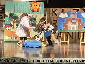 Scena teatralna. Dwie osoby w kostiumach interagują z rekwizytem na środku sceny. Jedna z nich ma strój białego ptaka/anioła i żółte buty. Tło stanowi kolorowa, bajkowa scenografia przedstawiająca las, słońce i zamek. U dołu sceny częściowo widoczny napis "PREZENTACJA ŻYCIA TWÓRCZEGO OSÓB NIEPEŁNOSP...".