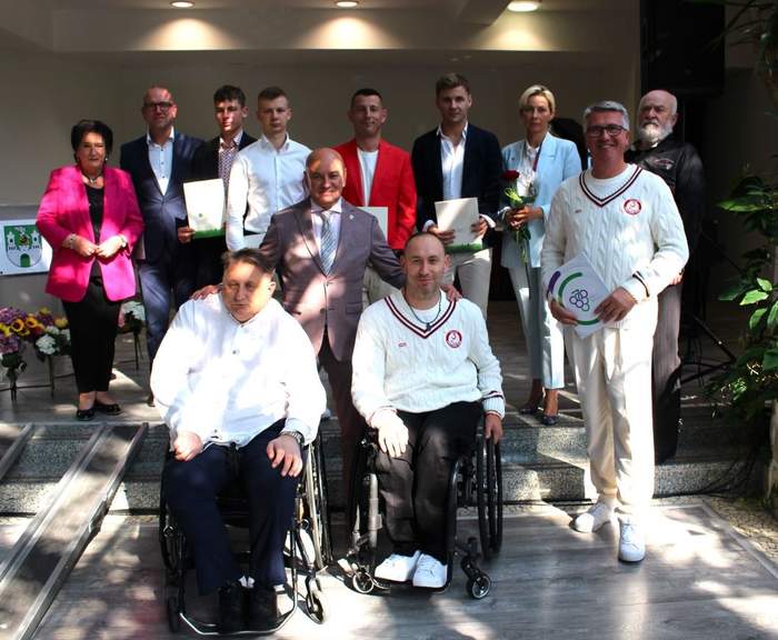 Pokaż zdjęcie: Zawodnicy, trenerzy i działacze stoją obok siebie w pamiątkowym zdjęciu, są osoby na wózkach, są ubrani na sportowo, niektórzy galowo