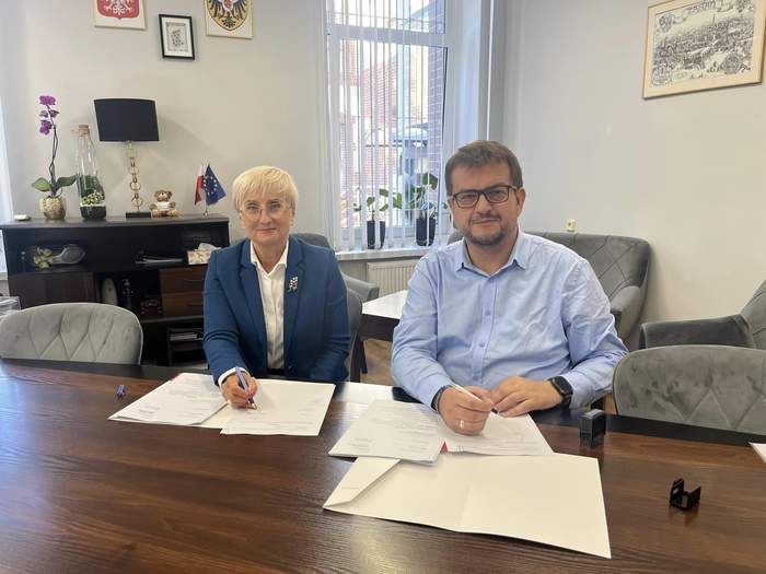 Pokaż zdjęcie: Kobieta i Mężczyzna siedzą przy biurku, są uśmiechnięci, ubrani na galowo, na biurku są podpisane umowy