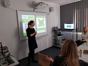 Pokaż zdjęcie: W centralnej części slajdu widać kobietę w czarnym stroju, stojącą przy tablicy projektowej i mówiącą do grupy. W tle jest wyświetlana prezentacja na tablicy. Po prawej stronie sali stoi wózek ze sprzętem i monitor, a przy oknie są zasłony. Na pierwszym planie widać siedzącą osobę z jasnymi włosami, od tyłu, zwróconą w stronę prowadzącej.