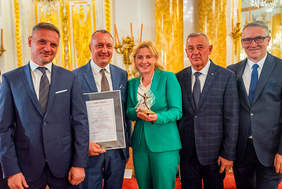 Pokaż zdjęcie: Do pamiatkowego zdjęcia pozuje 5 osób, 4 mężczyzn w garniturach oraz kobieta ubrana w jasnozielony garnitur. Kobieta trzyma w ręku statuetkę lodołamacza, obok niej mężczyzna trzyma w ręku dyplom.
