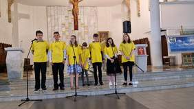 Pokaż zdjęcie: Trzej chłopcy i trzy dziewczynki stoją przed ołtarzem ubrani w żółte koszulki przed nimi stoją mikrofony.