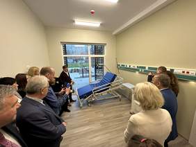 Pokaż zdjęcie: W pomieszczeniu stoją ludzie i obserwują prezentację łóżka podnoszonego elektrycznie. Osoba obok łóżka w czarnym swetrze i białej koszuli trzyma pilota. Podłoga wykonana z kafli we wzorze drewnianym jasne ciepłe ściany.