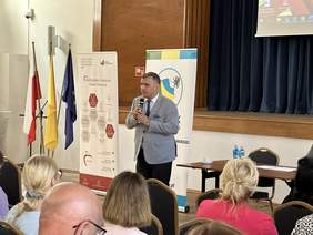 Pokaż zdjęcie: Mężczyzna w jasnej marynarce białej koszuli, trzyma mikrofon i mówi. Za nim stoją dwa banery i flagi w tym UE i Polska. Przed nim siedzą ludzie i słuchają. W tle kurtyna ze sceny.