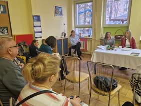 Pokaż zdjęcie: Zdjęcie z tyłu sali, na krzesełkach siedza rodzice, na tle okien stoi stół prezydialny przy którym siedzą trzy kobiety.