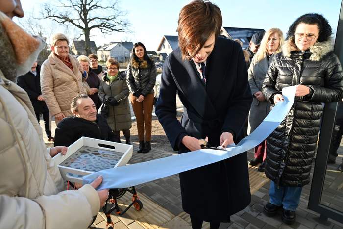 Pokaż zdjęcie: Kobieta trzyma w ręce nożyczki, którymi przecina błękitną wstęgę. W pobliżu znajduje się grupa kilkunastu osób. W tle widoczne są budynki mieszkalne oraz drzewa pozbawione liści.