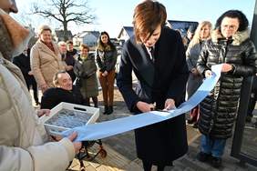 Pokaż zdjęcie: Kobieta trzyma w ręce nożyczki, którymi przecina błękitną wstęgę. W pobliżu znajduje się grupa kilkunastu osób. W tle widoczne są budynki mieszkalne oraz drzewa pozbawione liści.