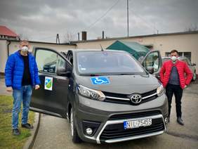 Pokaż zdjęcie: Na pierwszy plan wysuwa się szara toyota oznakowana na przedniej szybie piktogramem: dostępny dla osób z niepełnosprawnością, a na drzwiach herbem Gminy Iława.  Z lewej strony pojazdu stoi wysoki blondyn ubrany w niebieską kurtkę, jeansy i czarne