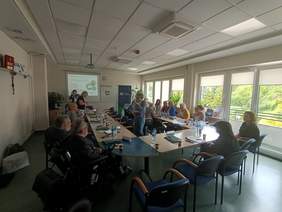 Pokaż zdjęcie: Kilkanaście osób siedzi przy dużym stole ustawionym w podkowę. Za nimi wyświetlona prezentacja oraz baner ZUS.