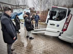 Pokaż zdjęcie: Operator kamery i dziennikarze stoją u tyłu samochodu. Kierowca furgonetki wysunął podjazd do wprowadzania wózków. Resztę osprzętu zasłaniają drzwi. 