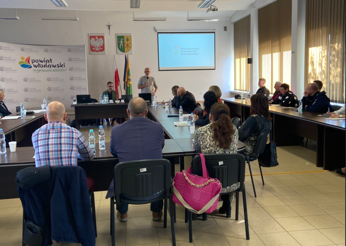 Pokaż zdjęcie: Sala konferencyjna z ekranem na środku, przy stołach siedzą uczestnicy konferencji