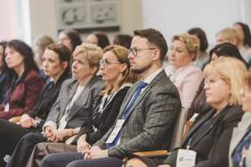 Pokaż zdjęcie: Zdjęcie typu portret zbiorowy z profilu. Na pierwszym planie widać mężczyznę w okularach i kraciastej marynarce, z niebieskim krawatem i identyfikatorem z napisem "GOŚĆ". Obok niego, w tym samym rzędzie, siedzą kobiety skupione na tym, co dzieje się przed nimi. Tło jest lekko rozmyte, co podkreśla koncentrację osób na pierwszym planie. Oświetlenie jest miękkie i naturalne.